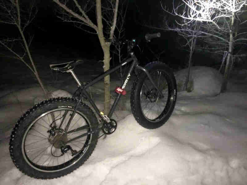 Right side view of a black Surly fat bike, parked in the snow against a tree, at night