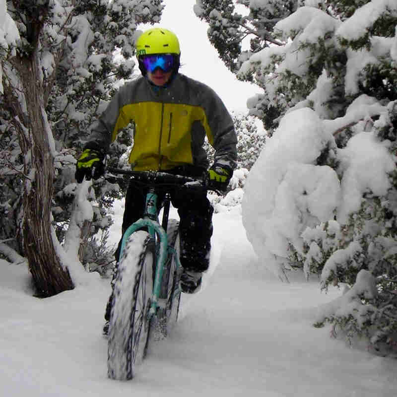 Front view of a cyclists on a mint Surly fat bike, riding through a narrow, deep snow, trail, in between trees