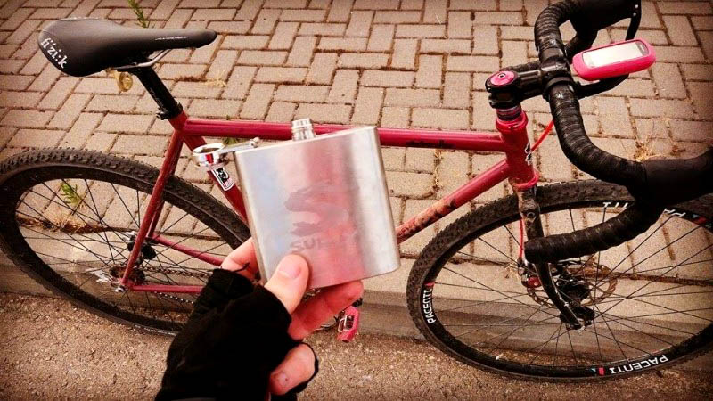 Downward view of a hand holding a Surly flask above a Surly Steamroller bike, red, parked against a street curb
