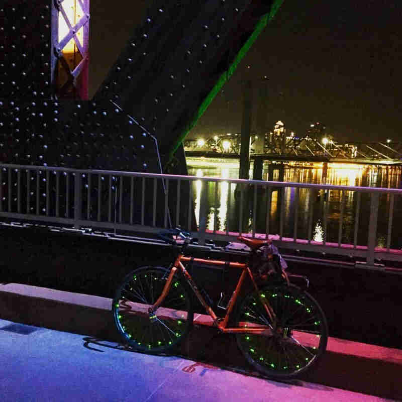 Left side of view of a Surly bike, orange, parked along the side of a bridge, with a city in the background at night
