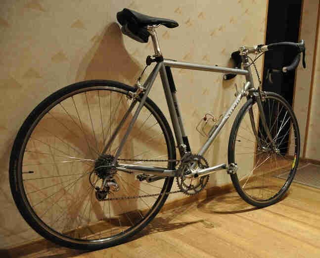 Right side view of a silver Surly bike, leaning against a wall, in a room with wood floors