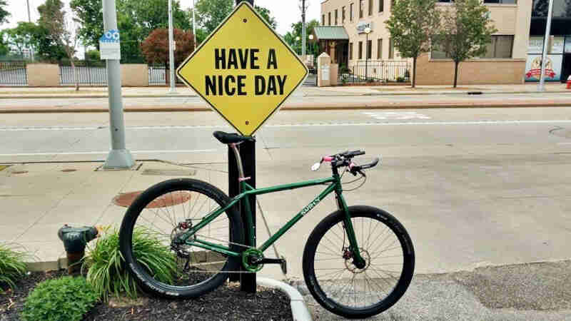 Right view of a green Surly bike, parked next to a sidewalk, in front of a Have a Nice Day sign