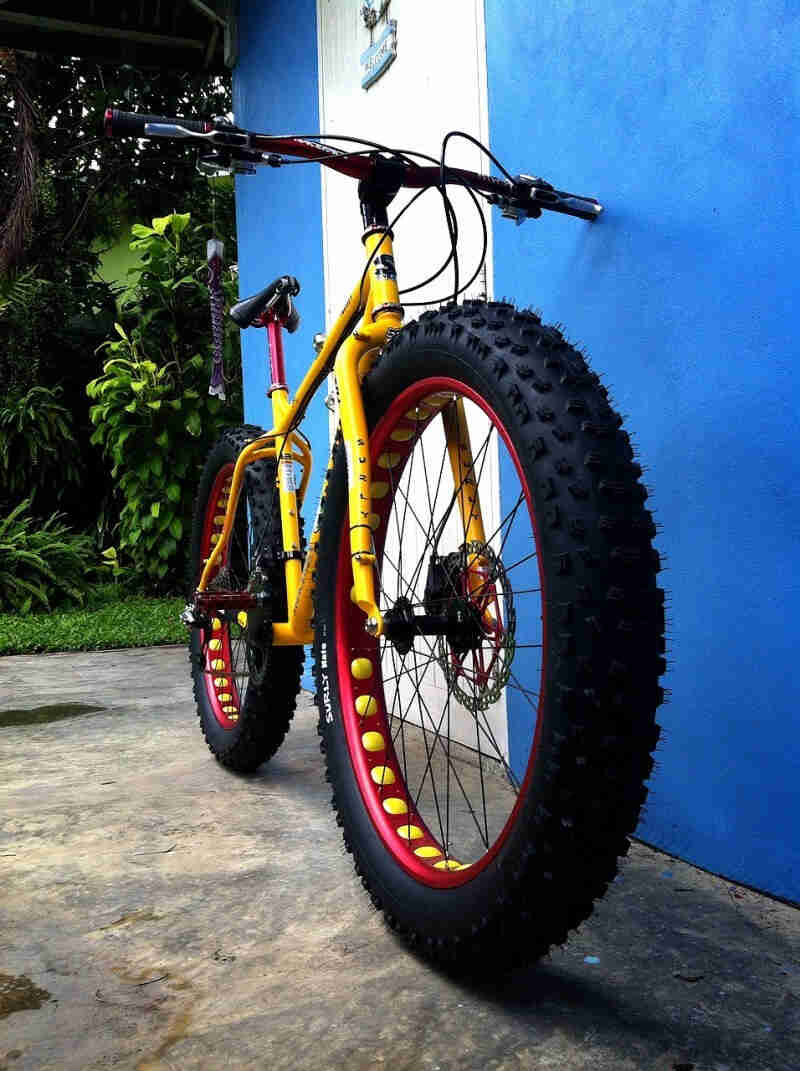 Front, right side view of a yellow Surly fat bike with red rims, leaning a blue wall with a white door, on a cement slab