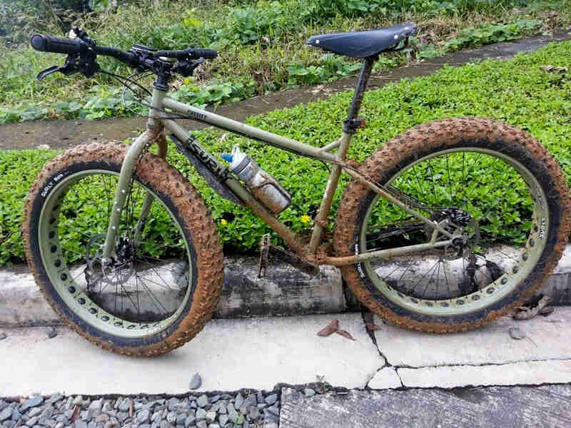 Left side view of a muddy, olive Surly Pugsley fat bike, parked against a street curb