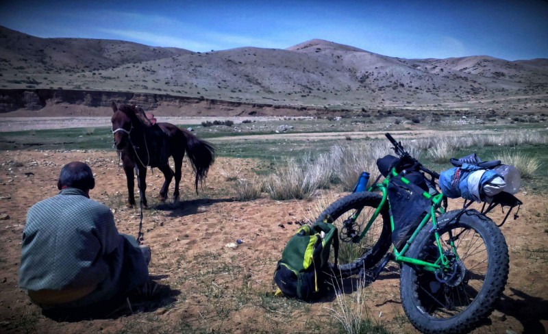 Rear view of a person sitting in sand next to a Surly fat bike, facing a horse, with mountain hills in the background