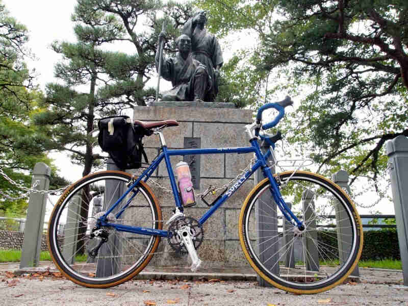 Right side view of a blue Surly Long Haul Trucker bike, parked in front of a statue with stone base, and trees behind it