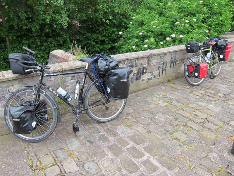 Left side view of a black Surly bike, loaded with gear, on a sidewalk, leaning a short, brick wall with shrubs behind it