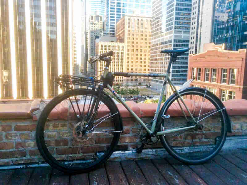Left side view of a green Surly bike, park against the ledge of a rooftop, with city buildings in the background