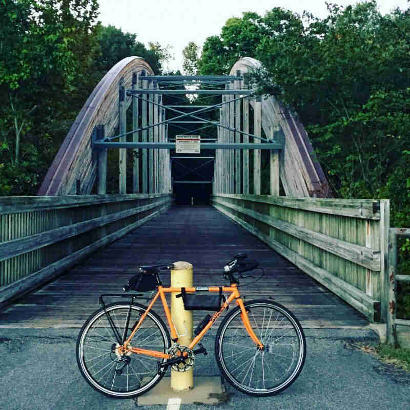 Right profile of a Surly bike, orange, parked across a paved trail, in front of a  bike bridge that leads into a forest