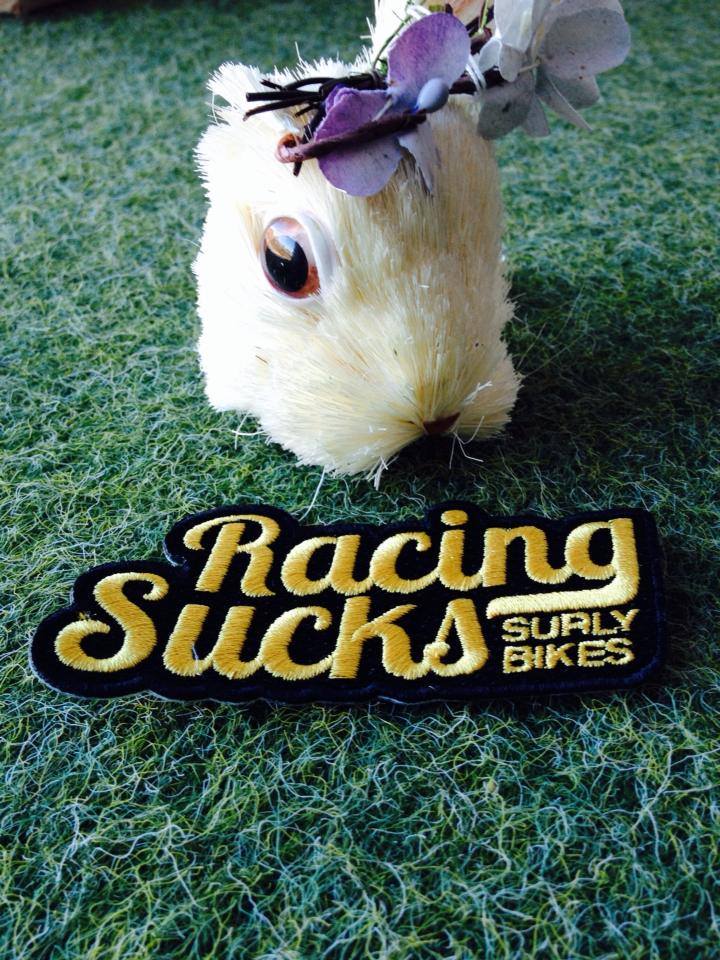 A Surly Bikes Racing Sucks patch, in front of the head of a toy gopher, laying on artificial grass