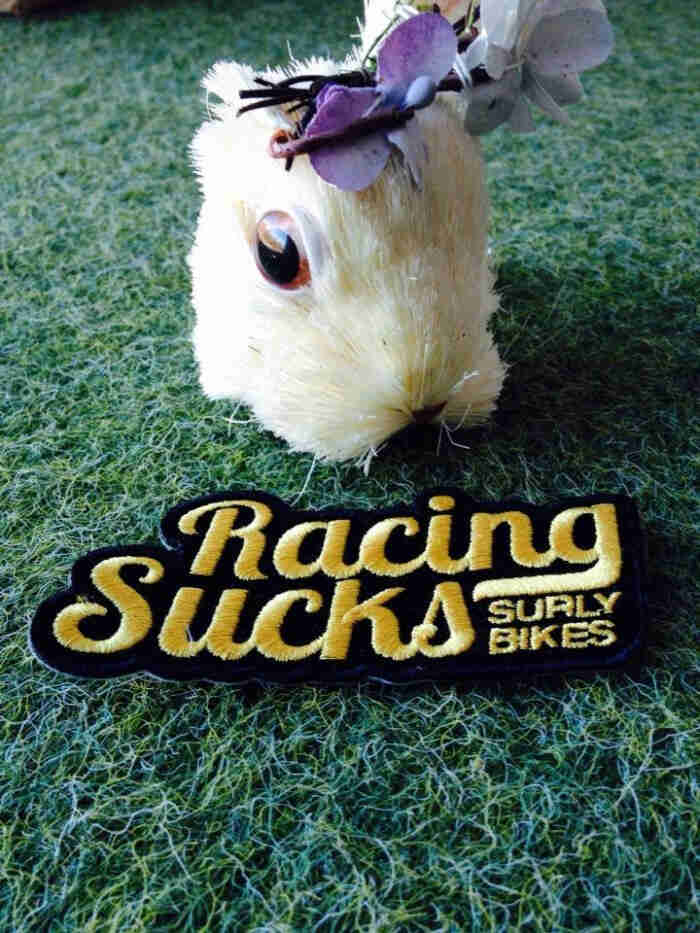 A Surly Bikes, Racing Sucks patch, in front of the head of a toy gopher, laying on artificial grass