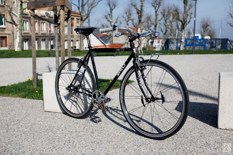 Right side view of a black Surly Cross bike, leaning on a cement block on a courtyard, with trees and buildings behind