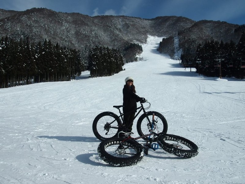 Right side view of a cyclist on a Surly fat bike, next to a fat bike laying down, on a snowy ski hill in the mountains