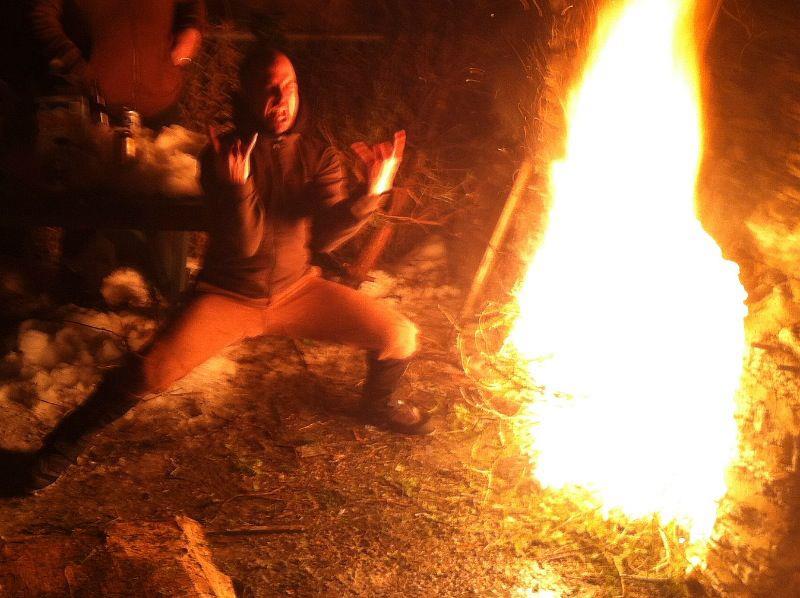 Front view of a person bending down with their right left out straight, on the side of a campfire at night