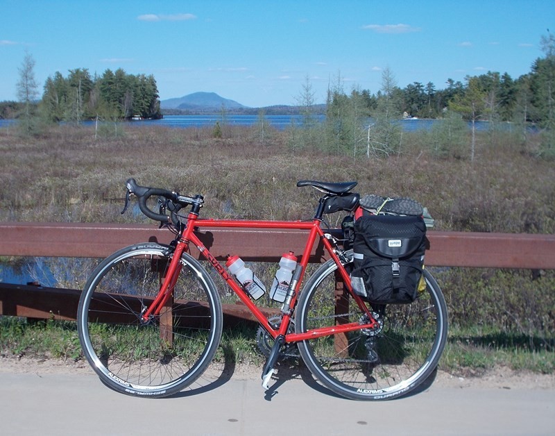 Left side view of a red Surly bike with rear packs, leaning on a railing, with wetlands, a lake and mountains behind it