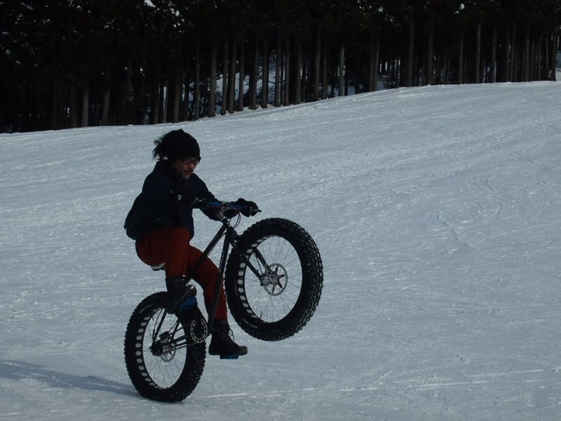 Front, right side view of a cyclist, riding a wheelie on a Surly fat bike, across the bottom of a snow covered field