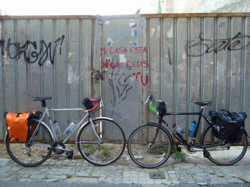 Side view of 2 Surly bikes with gear, parked on a brick sidewalk while facing each other, in front of a steel wall