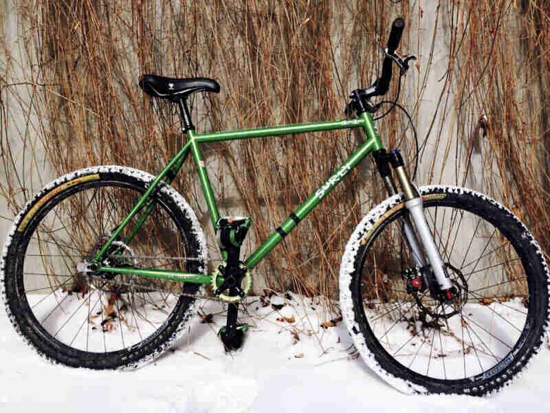 Right side view of a green Surly 1x1 bike, parked in snow and leaning against a cement wall with weeds handing down