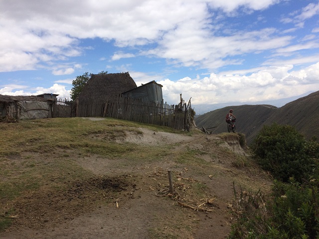 Rear view of a cyclist riding on a dirt trail, next to a mountain homestead, with hills in the background