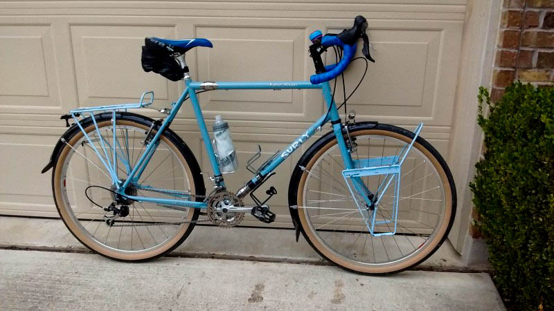 Left side view of a turquoise Surly bike, on a driveway, leaning on a garage door 
