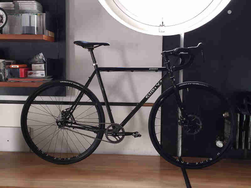 Right side view of a Surly Straggler bike, parked on a wood floor, against a wall in a room