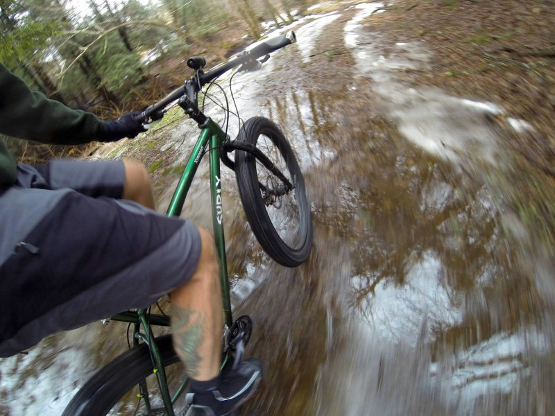 Downward right side rear view of a cyclist riding a Surly Krampus bike, green, down a muddy trail in the woods