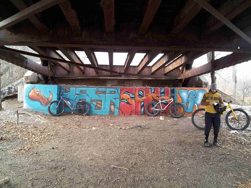 Front view of a cyclist standing under the steel beams of a bridge, with their yellow Surly fat bike behind them