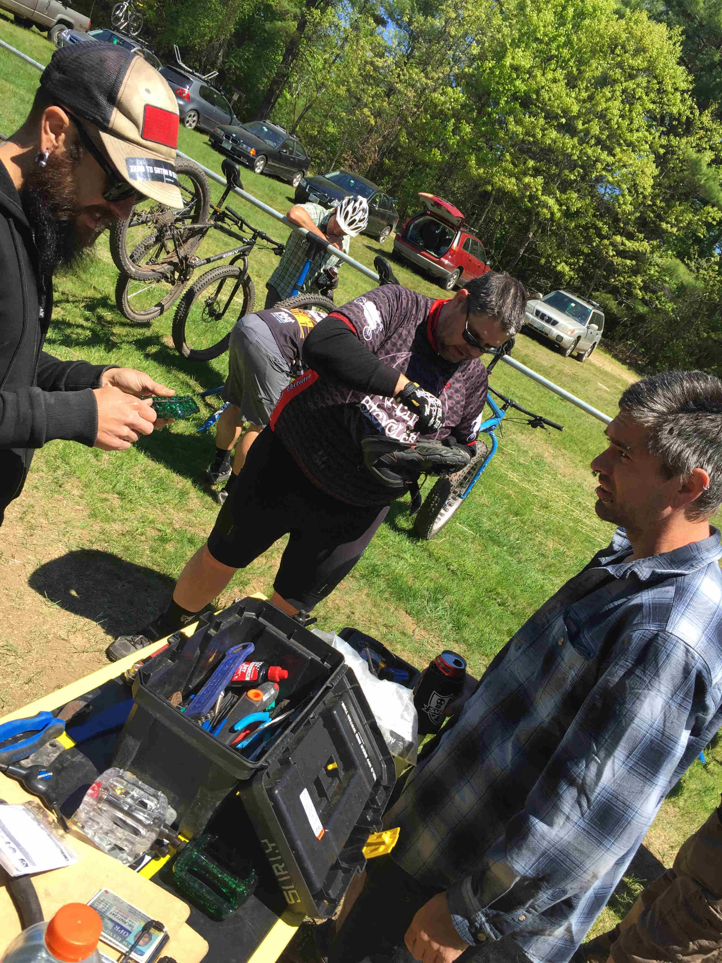 Tilted view of 3 people standing around a table with an open tool box, with cyclists and bikes in the background