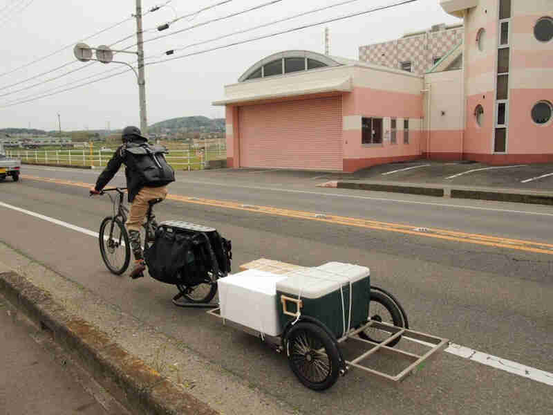 Rear of a cyclist riding down the side of a roadway on a bike, with a trailer hauling coolers, hitched behind