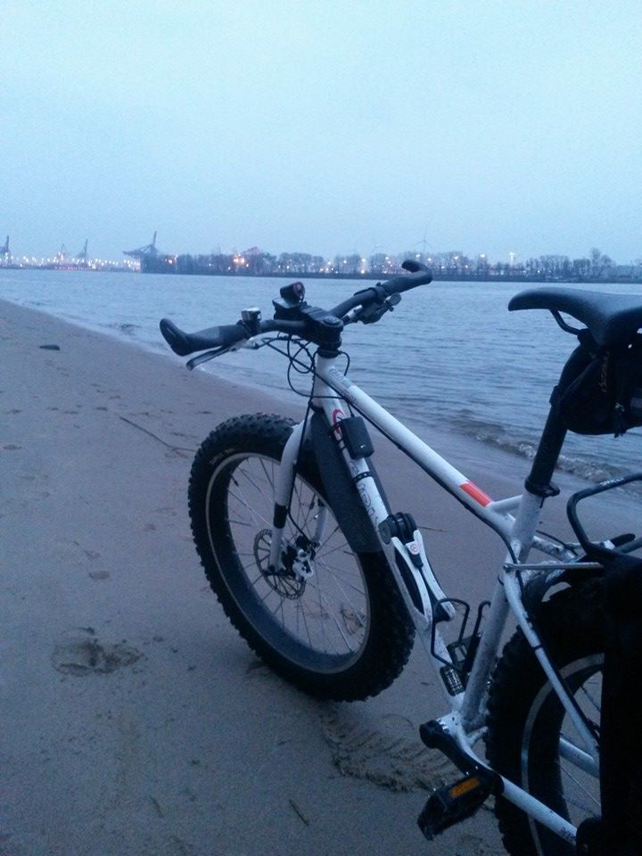 Rear, left side view of a white Surly fat bike, parked along a sandy shore, next to a waterway at dusk on a hazy day