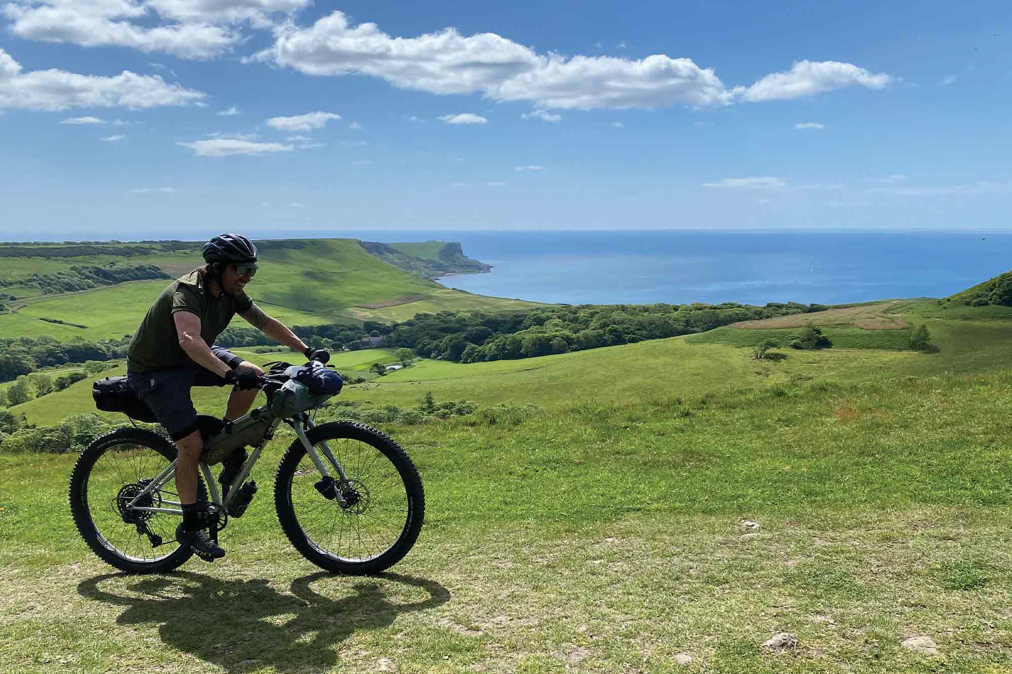 Cyclist w/ bikepacking bags riding grassy hills on sunny day with shoreline in distance