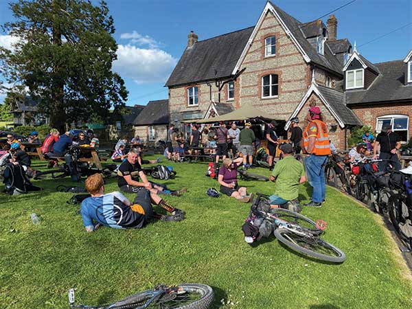 People gathered outside pub, sitting on tables and grassy, bikes parked