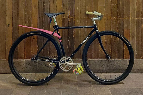Black Surly Steamroller with cruiser bar and pink rear fender leaning against fence outside