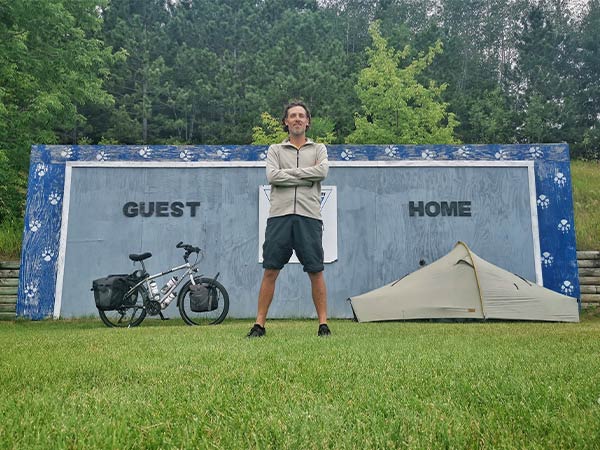 Yann standing in front of 'Guest home' sign with tent set-up and bike parked on grass lawn