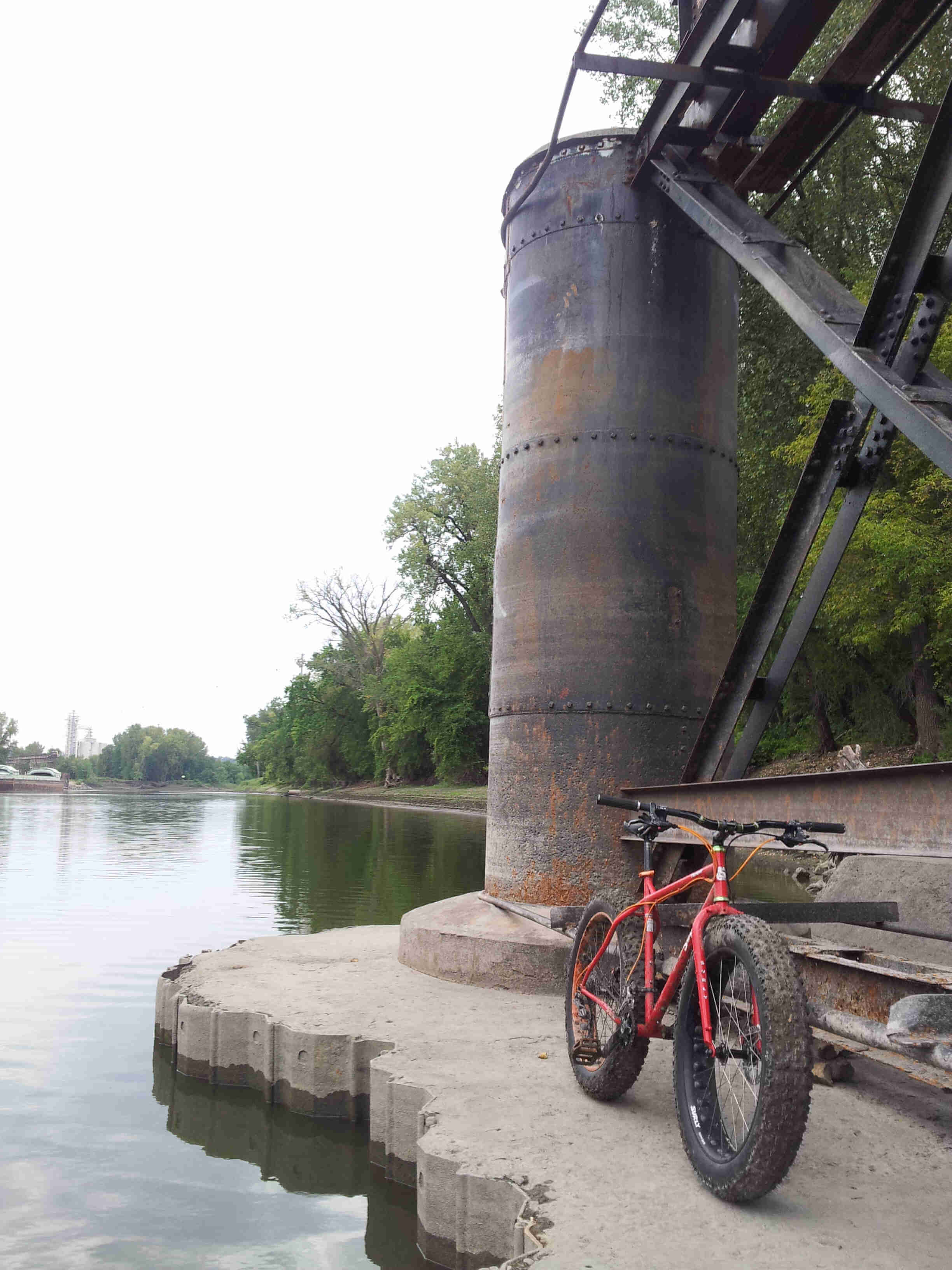Front, right side view of a red Surly Moonlander fat bike, parked on a platform with a river bridge post on it