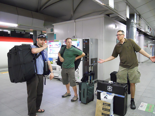 Front view of 3 people standing with their luggage, inside of a train terminal building