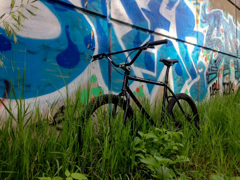 Front, left view of a black Surly 1x1 bike, parked in tall weeds, against a graffiti covered wall