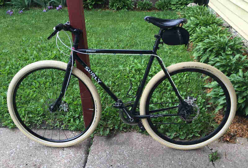 Left side view of a black Surly bike on a sidewalk, next to a yard with green grass