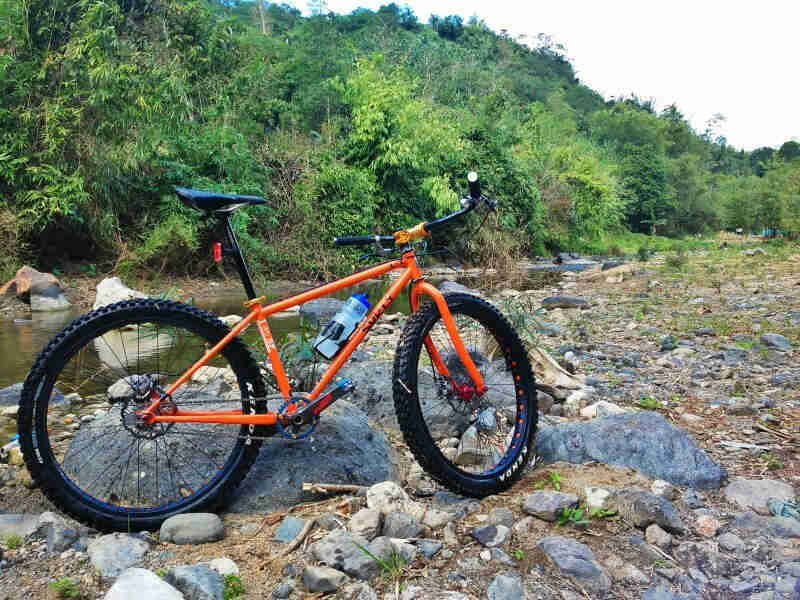Right side view of an orange Surly 1x1 bike, parked on a rocky shore next to a river, with tree covered hills behind it