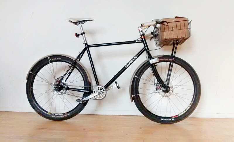 Right side view of a black Surly 1x1 bike with front basket, leaning against a white wall