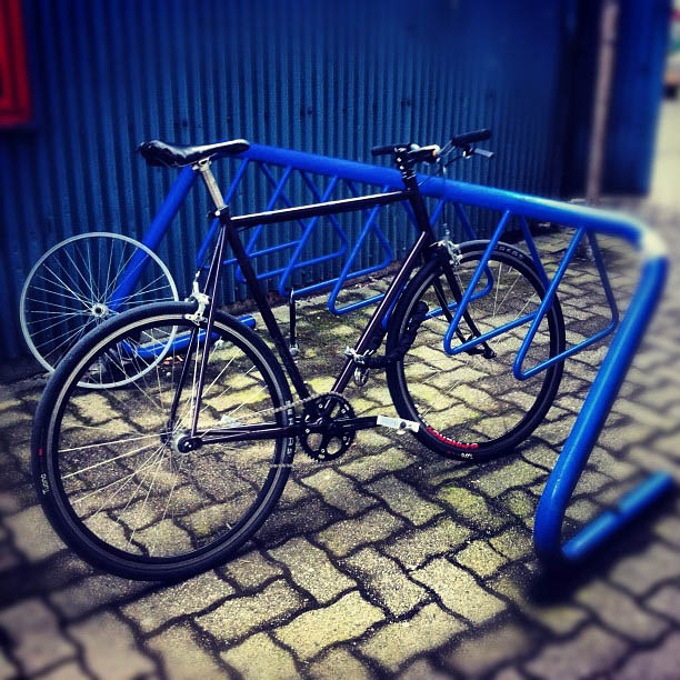 Right side view of a black bike, parked in a blue bike rack against a blue wall