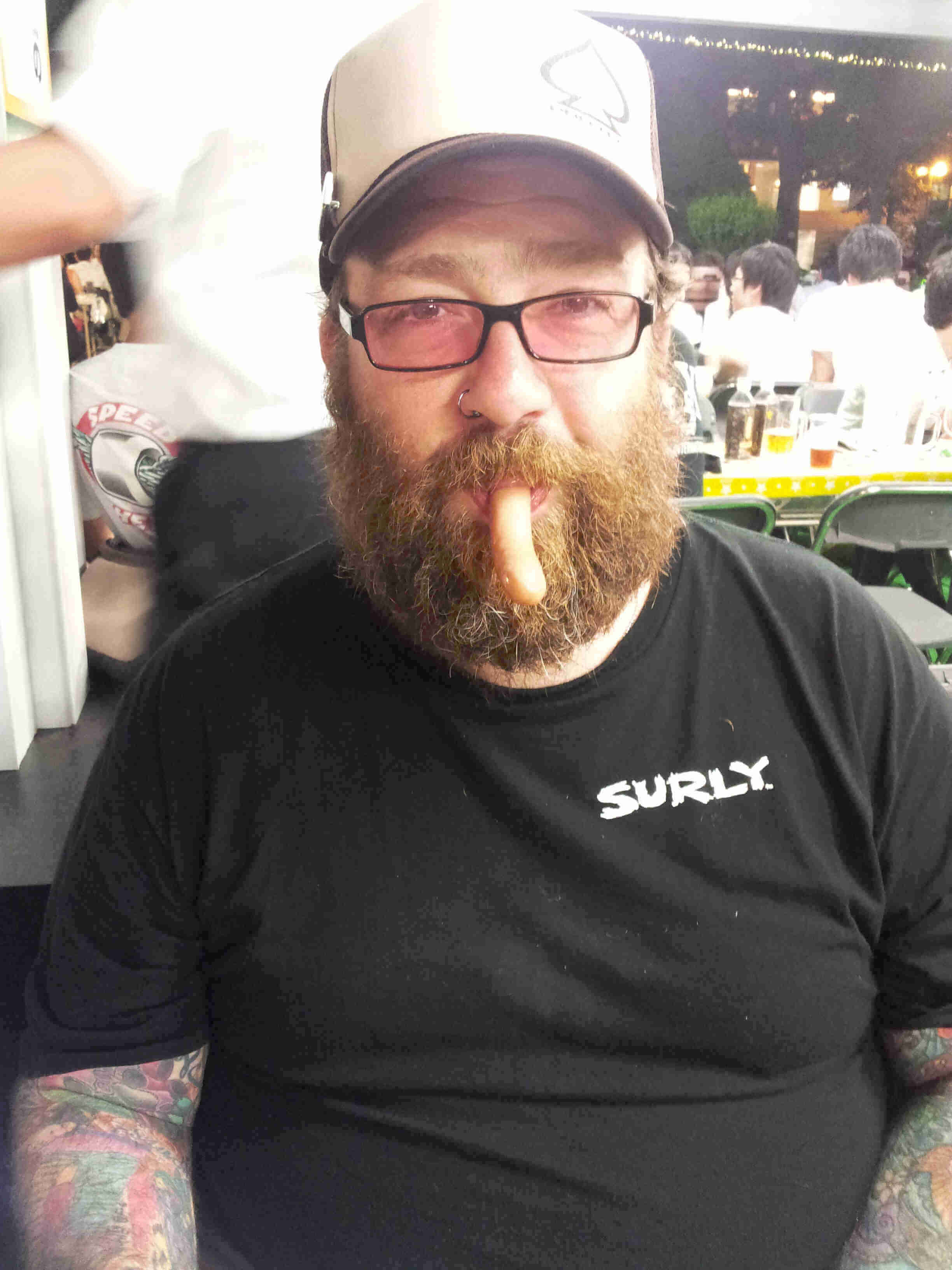 Front, waist up view of a person with a beard, and piece of food hanging from their mouth, wearing a black Surly t-shirt