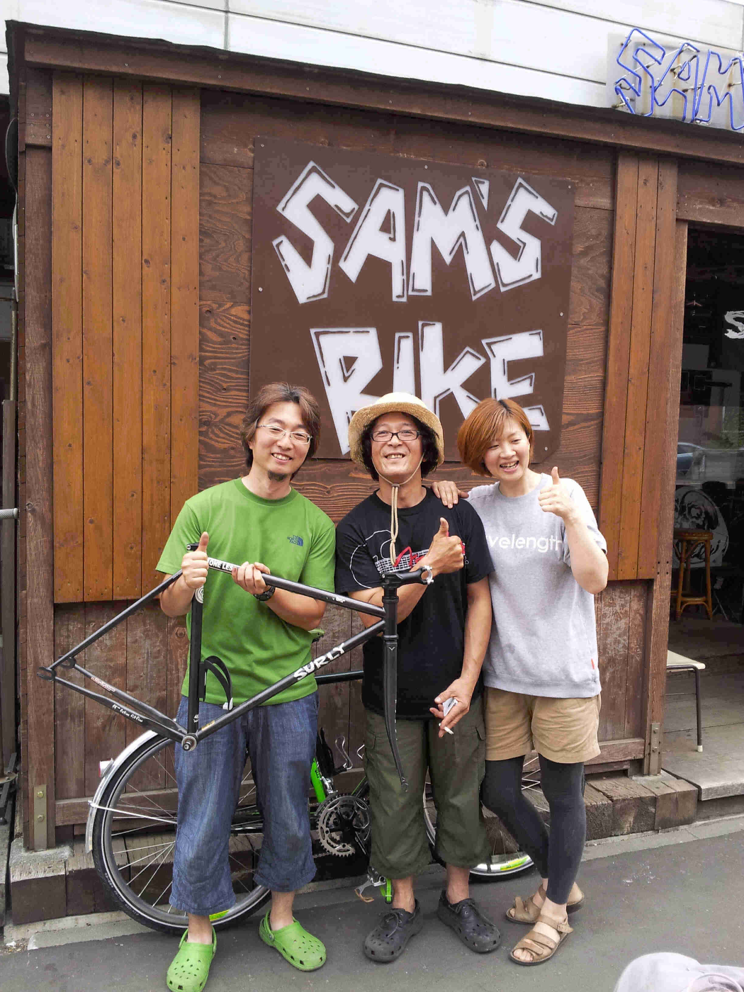 Front view of a person, holding up a Surly bike frameset, standing with 2 other people, outside of Sam's Bike shop