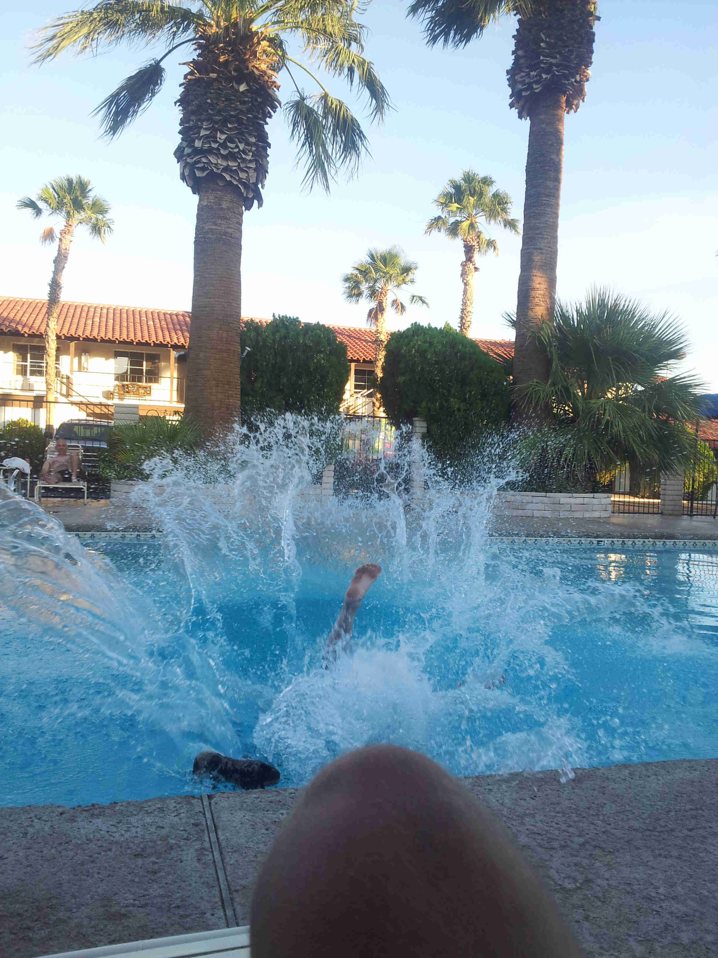 A person splashing into an outdoor pool with palm trees around it, with a motel in the background