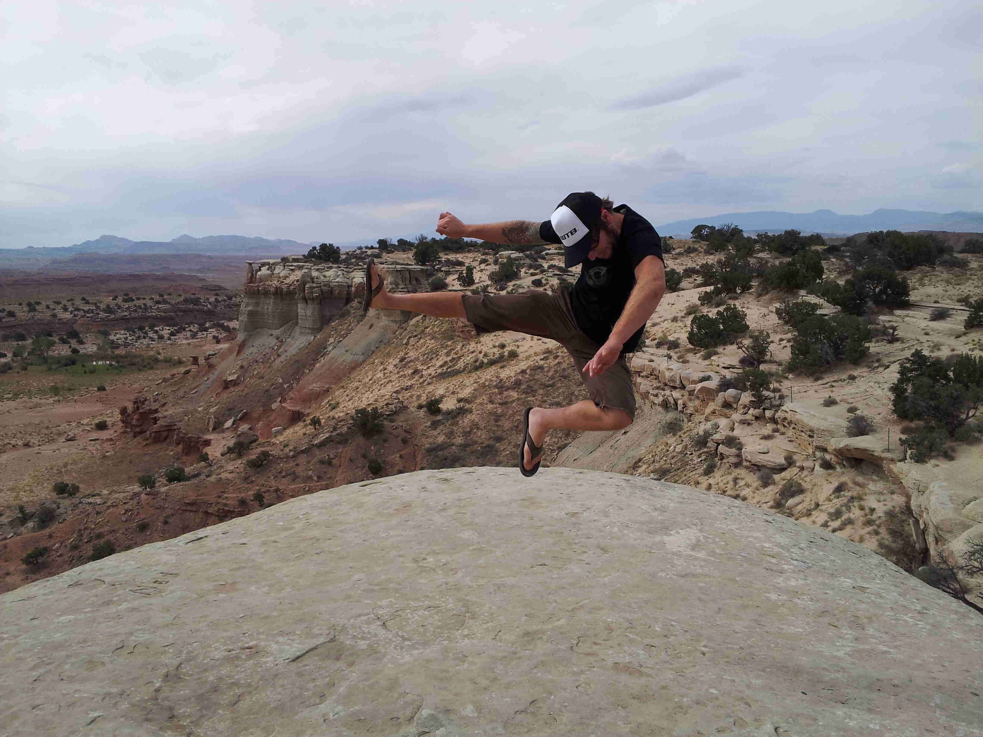 Front view of a person leaping in the air, on a large, smooth boulder, above a desert canyon that's behind them