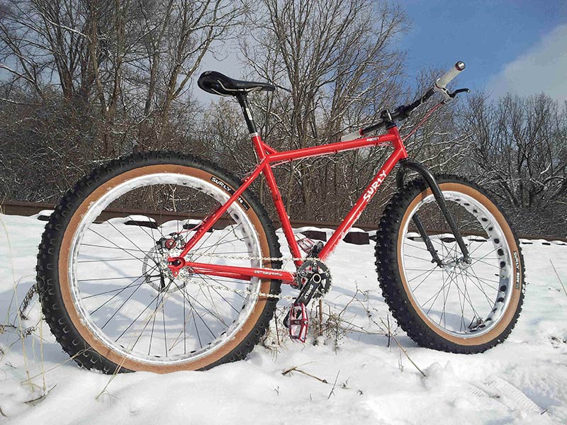 Right side view of a red Surly Pugsley fat bike, parked in snow along a small, snow covered hill with train tracks above