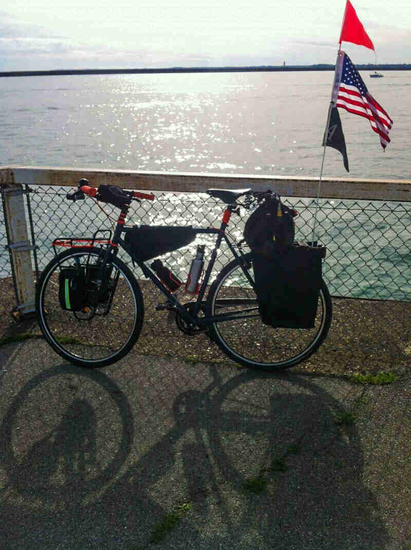 Left side view of a bike with gear packs, and a vertical flagstick with the US flag, leaning on a fence next to a lake