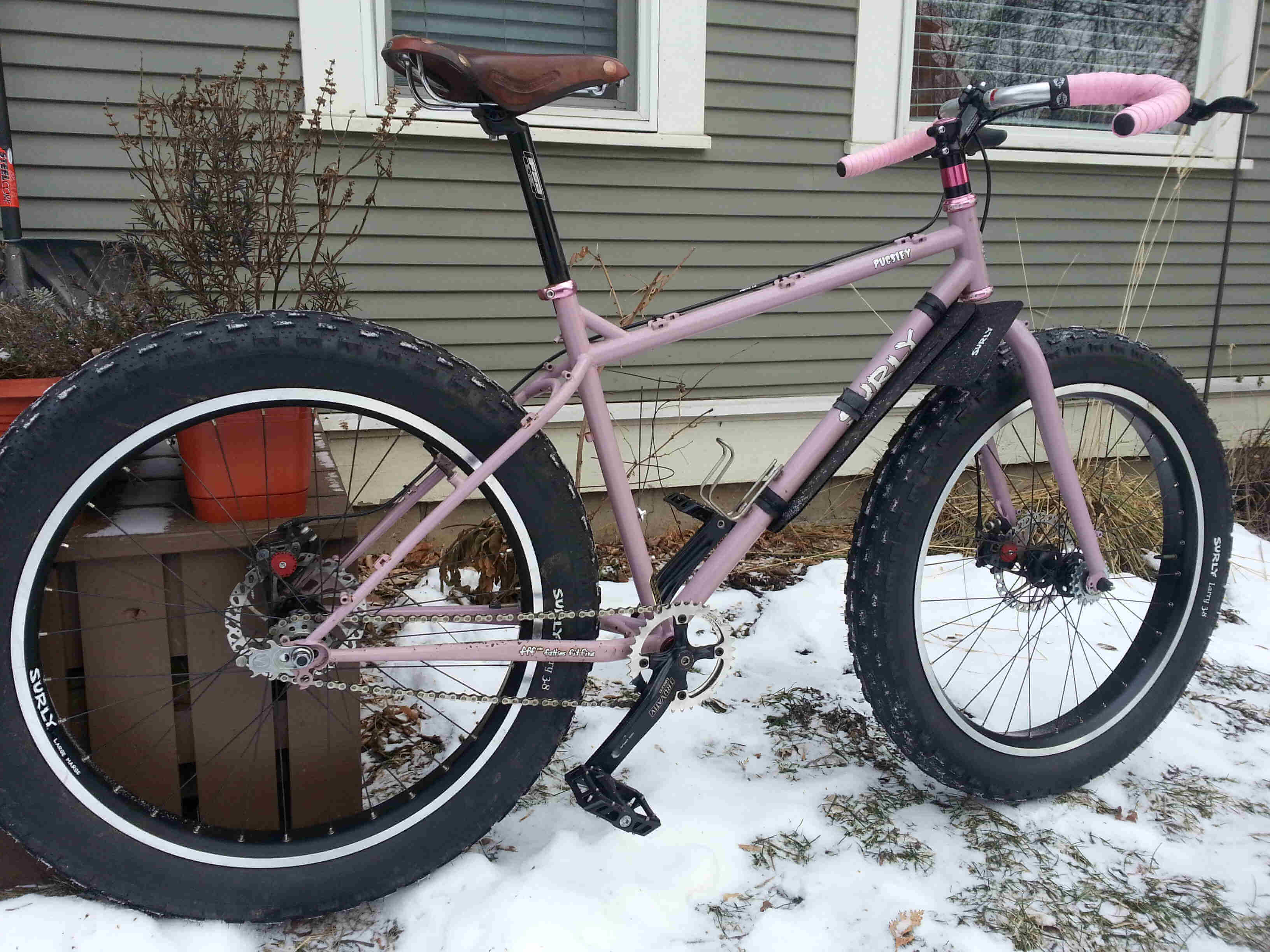 Right side view of a pink Surly Pugsley fat bike, parked on snow, near the side of a gray house