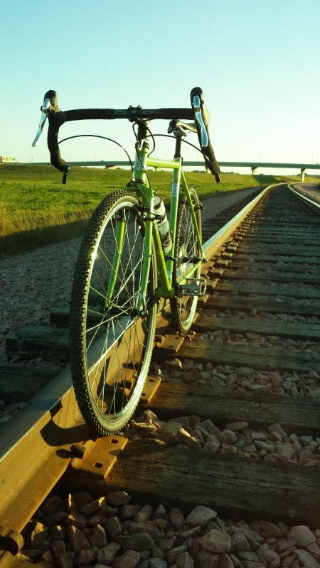 Front view of a green bike, parked along one of the rails on train tracks, with a field and bridge in the background