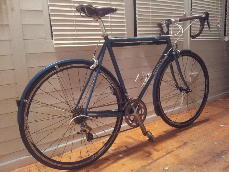 Right side view of a blue Surly Pacer bike, leaning against a wall with blinds on the window, in a room with wood floors
